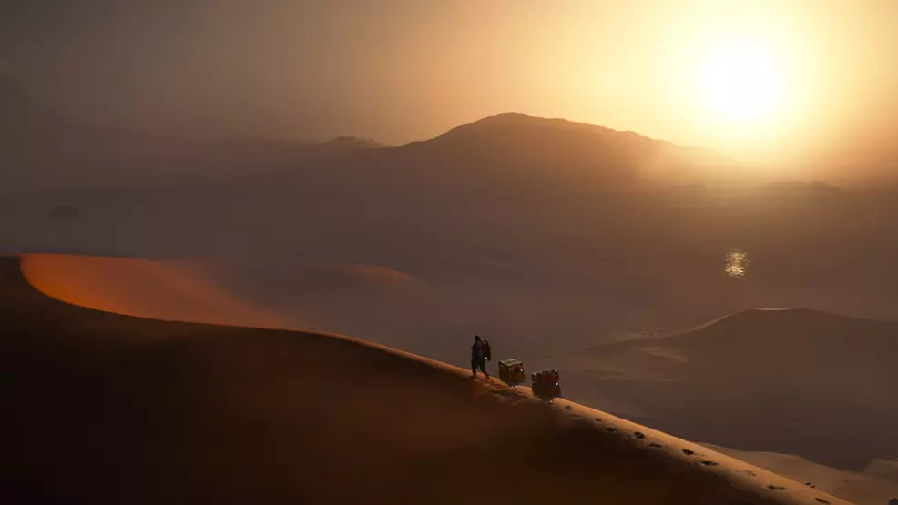 The Sam trek with two cargo carriers in the desert, which tags behind it in Death Stranding 2.